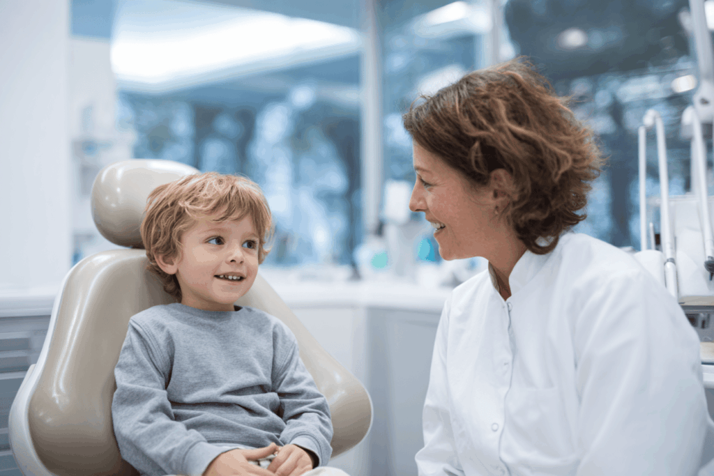 Jeune enfant d’environ 4 ans assis dans un fauteuil de dentiste, regard curieux et rassuré, dentiste souriant en blouse blanche expliquant un instrument dentaire, cabinet lumineux et moderne, couleurs douces, ambiance bienveillante, parent présent à côté de l’enfant, photographie réaliste, lumière naturelle, style éditorial médical.