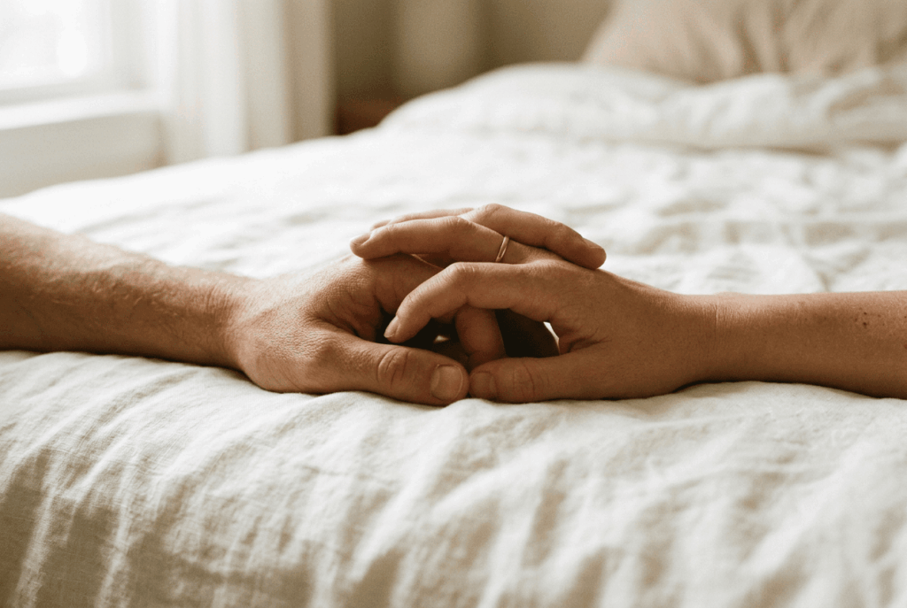 Le câlin du matin couple sur le lit avec gros plan sur deux mains entrelacées posées sur drap blanc. Lumière douce et naturelle venant du côté gauche. Tons neutres et chauds — beige, ivoire, peau. Pas de bijoux voyants. L'image évoque la complicité, la tendresse matinale et la sécurité affective.