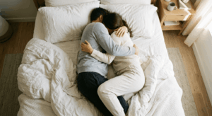 Vue plongeante prise depuis au-dessus du lit, couple allongé côte à côte, habillés en tenues décontractées et douces (blanc, beige, gris clair). Ils s'enlacent tendrement, visages non visibles — on voit seulement leurs corps blottis l'un contre l'autre, les bras entrelacés. Draps blancs froissés, lumière naturelle dorée du matin filtrant par une fenêtre hors cadre. Ambiance chaleureuse, intime et apaisante. Style photo lifestyle épuré.